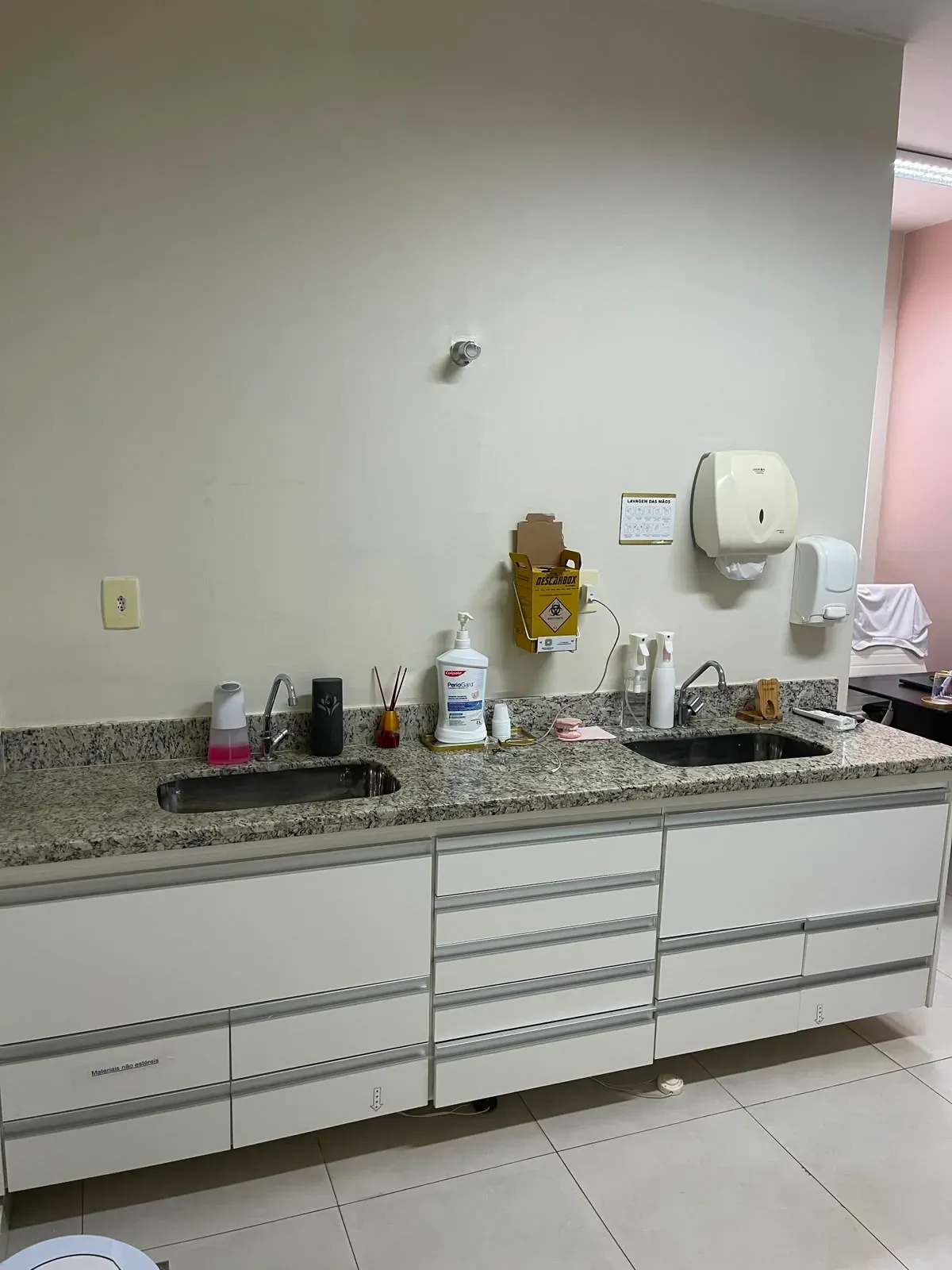 Bancada de granito com duas cubas, armários brancos e dispensers de higienização do consultório