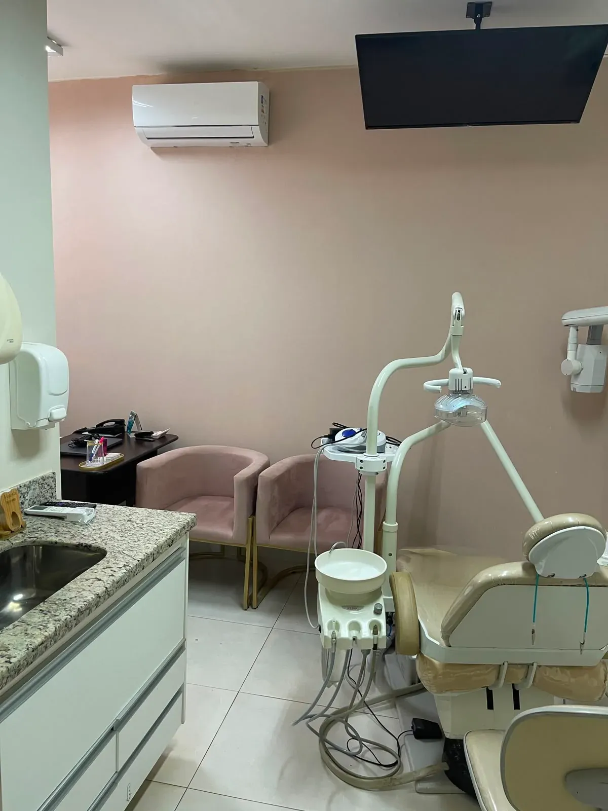 Consultório com cadeira odontológica, televisão no teto, ar-condicionado e poltronas ao lado da bancada